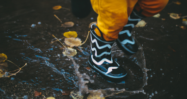A close-up of a child standing in a puddle. They are wearing yellow pants and blue rubber boots with white stripes on them.